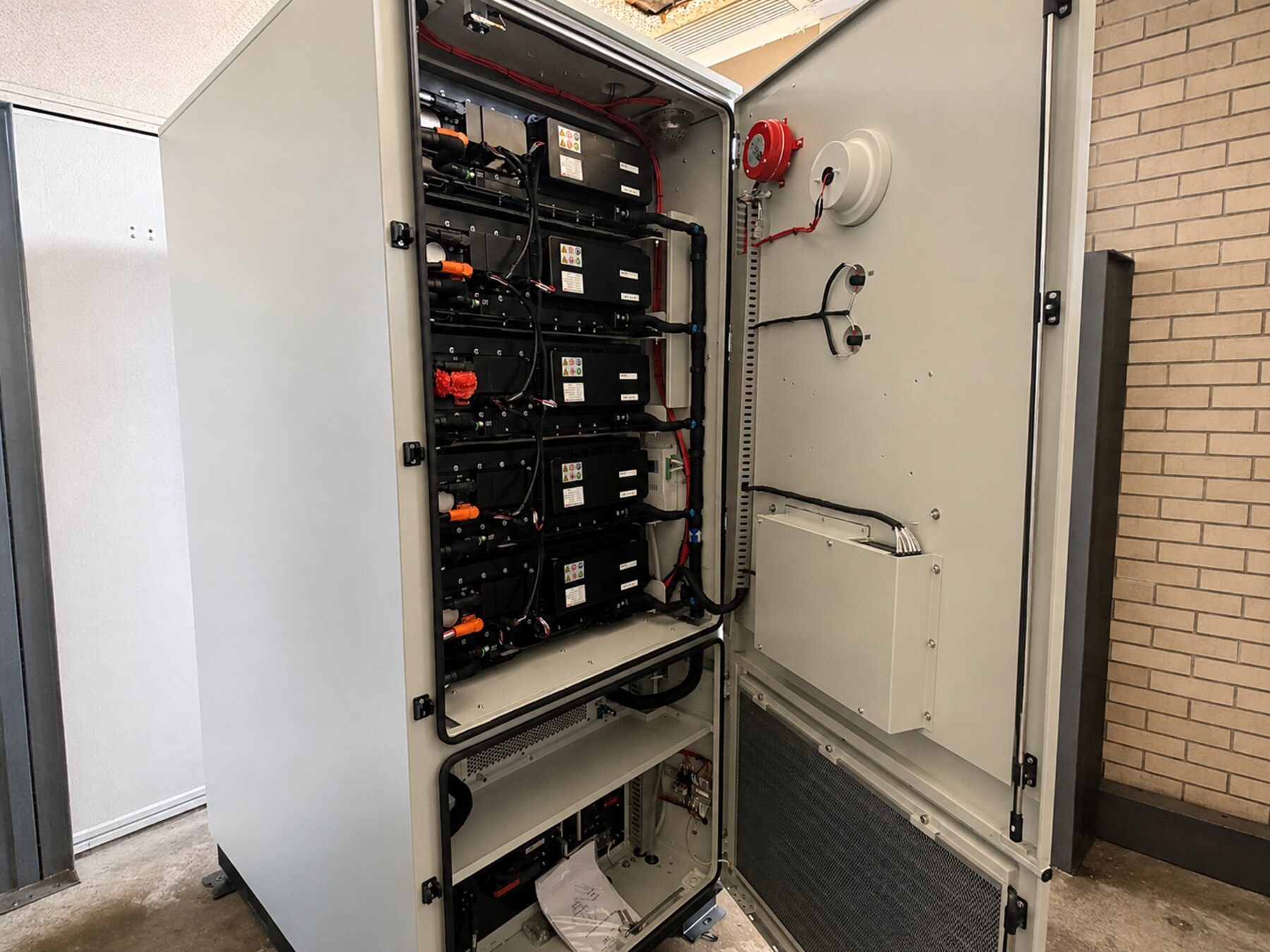 Ein Speicherschrank steht offen vor einer Ziegelwand: fünf Batteriemodule übereinander, die Leistungs- und Signalverkabelung am Türrahmen entlang geführt
