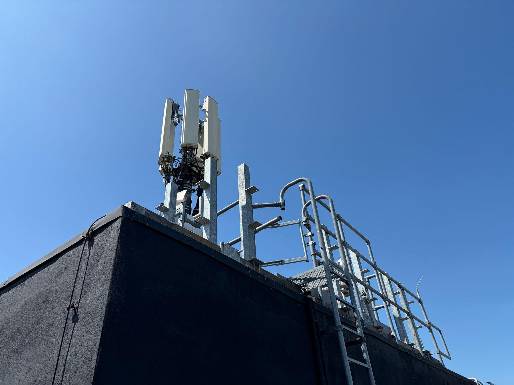 Antennenpaneele auf einem Mast an der Dachkante vor klarem Himmel, daneben Leiter und Geländer