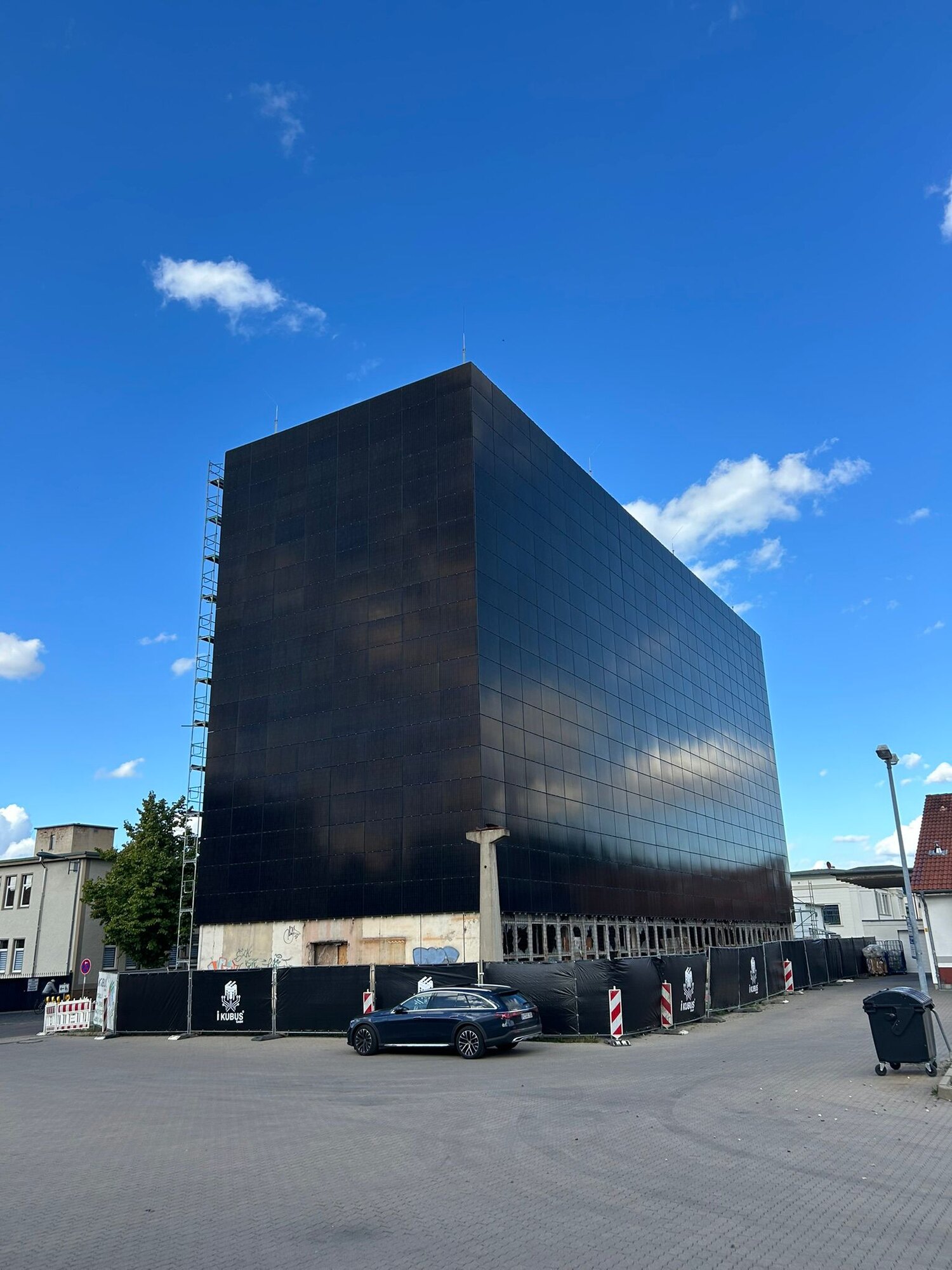 Der Kubus von der Straßenecke: das ganze photovoltaikverkleidete Gebäude, Bauzaun an seinem Fuß, davor ein geparktes Auto